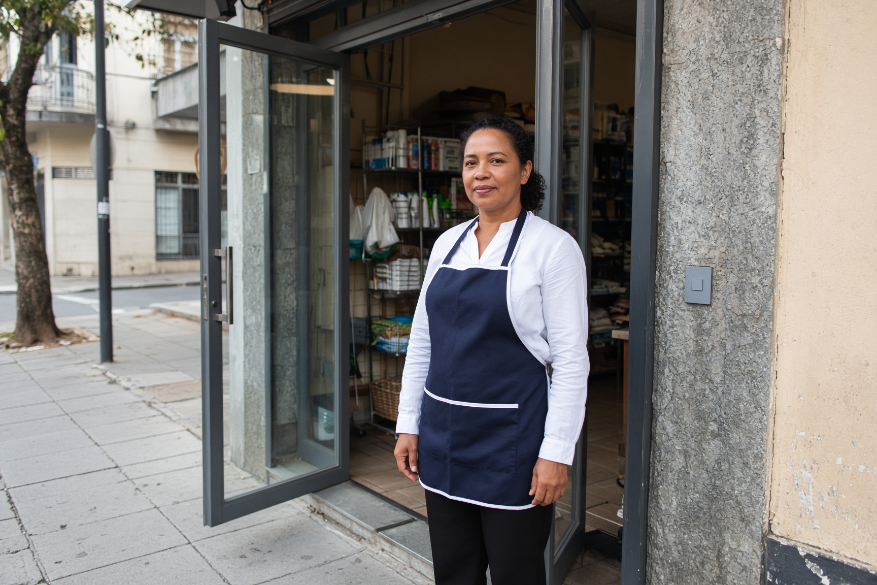 Profissional de limpeza, de avental azul e blusa branca, na porta de uma loja de rua